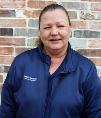 Middle-aged woman smiling in front of a brick wall, wearing a blue work jacket with text "Grace Veterinary Clinic" embroidered on it.