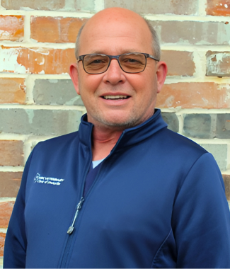 Bald middle-aged man in glasses smiling, wearing a blue jacket, against a brick wall.