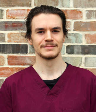 Man with a ponytail wearing a maroon scrub top standing in front of a brick wall.