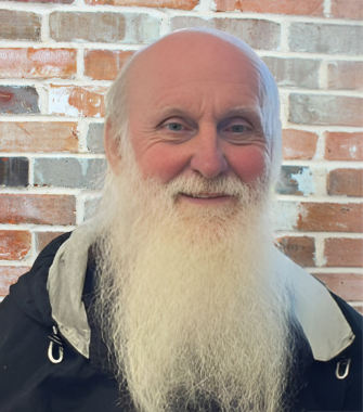 Elderly man with a long white beard smiling in front of a brick wall.