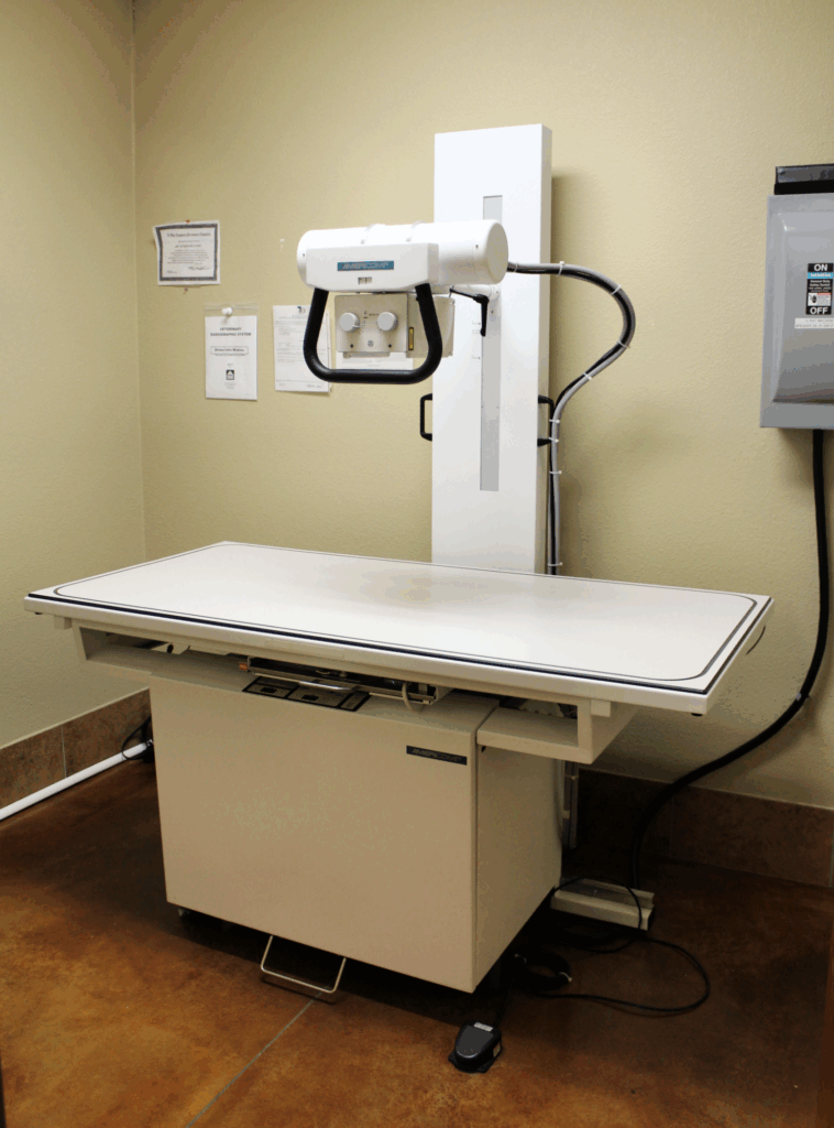 X-ray machine with adjustable table and overhead imaging tube in a medical room.