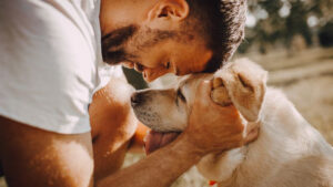 A man tenderly touches his forehead to his dog's head in a sunny outdoor setting.