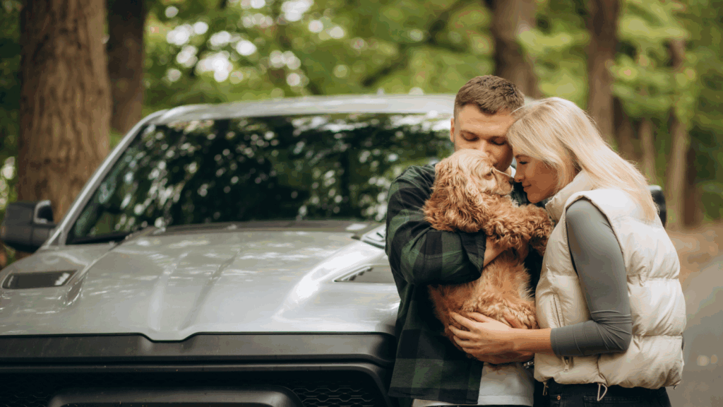 Couple embracing their dog near a car in a wooded area.